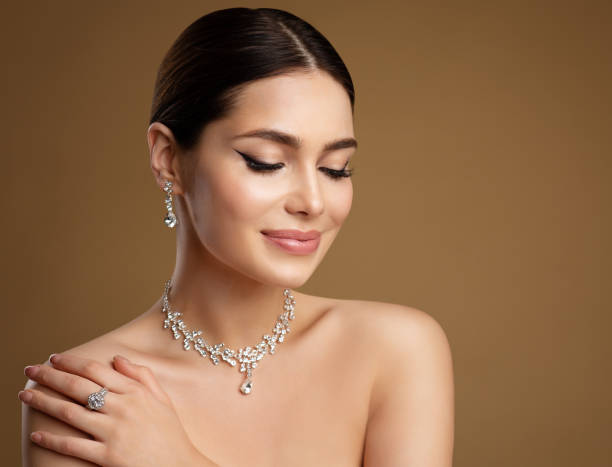 Beauty Model in Wedding Jewelry Set. Elegant Woman in Necklace with Earring and Ring. Beautiful Girl with perfect Eyeliner Make up and smooth Skin  looking down over Beige Background